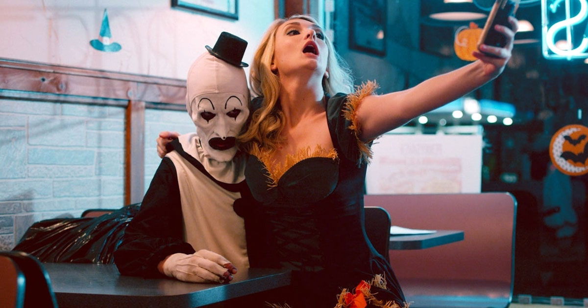 A scene from the Terrifier film series showing the Miles County Clown in a diner with a young woman sitting on his lap, using her mobile phone to take a selfie.